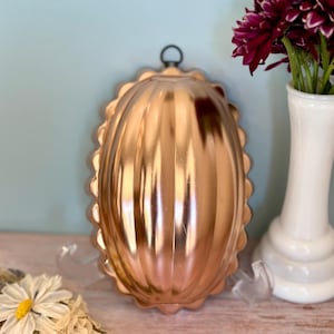 May include: A copper-colored, oval-shaped vintage jelly mold with a scalloped edge and a hanging loop. The mold is displayed on a wooden surface, next to a white vase with dark red flowers and a small bouquet of dried flowers.