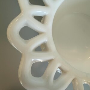 May include: Close-up of a white ceramic bowl with a decorative, openwork rim. The bowl has a smooth, glossy finish and a shallow interior. The rim features a repeating pattern of curved shapes with open spaces, creating a delicate, lace-like effect.