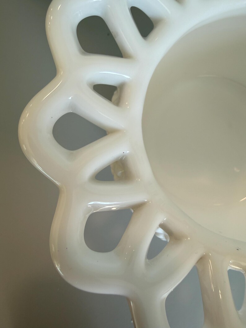 May include: Close-up of a white ceramic bowl with a decorative, openwork rim. The bowl has a smooth, glossy finish and a shallow interior. The rim features a repeating pattern of curved shapes with open spaces, creating a delicate, lace-like effect.