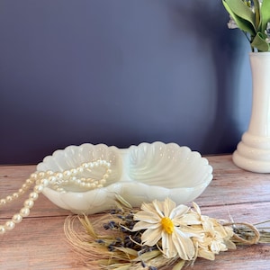 May include: A white milk glass double shell dish with a scalloped edge. A strand of cream-colored faux pearls rests inside one of the shell compartments. The dish is positioned near a small dried floral bouquet featuring white daisies and lavender.