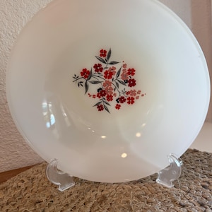 May include: A white, round serving bowl with a floral design in the center. The flowers are red and pink with black accents. The bowl is displayed on a clear plastic stand, resting on a woven mat.