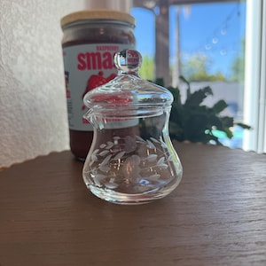 Op de afbeelding: Een heldere glazen creamer met een deksel, met geëtste bloemmotieven. De creamer heeft een kleine tuit en een ronde basis. Een pot frambozenjam staat op de achtergrond. De creamer staat op een bruin houten oppervlak.