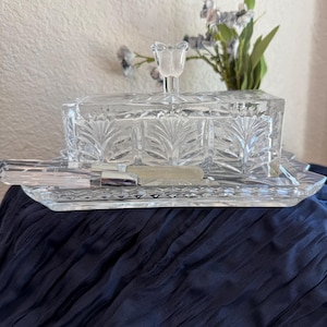 May include: A clear glass butter dish with a rectangular base and a matching lid featuring a floral pattern and a tulip-shaped handle. A clear glass butter knife with a silver-toned metal blade rests on the tray in front of the lid.