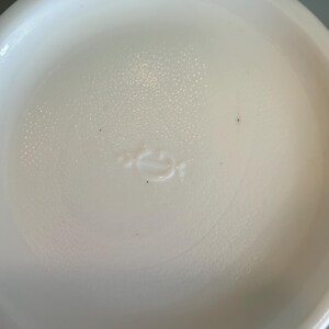 May include: A white, round, ceramic dish with a slightly textured surface. The dish has a shallow depth and a smooth, curved rim. A small, embossed design is visible in the center.