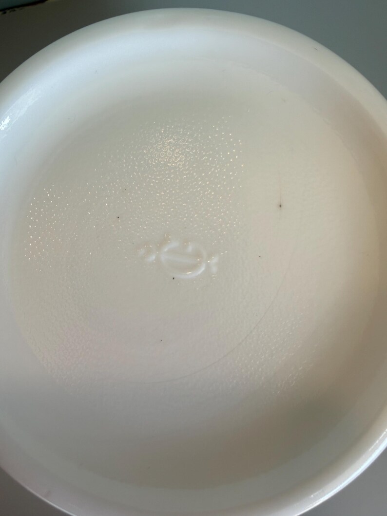 May include: A white, round, ceramic dish with a slightly textured surface. The dish has a shallow depth and a smooth, curved rim. A small, embossed design is visible in the center.
