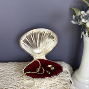 以下が含まれることがあります： 赤いベルベットの内装が施された、銀色の貝殻型のジュエリーボックス。ボックスには金のネックレスと2つの指輪が入っています。ボックスは白いレースの表面に置かれ、背景には白い花瓶に入った花があります。