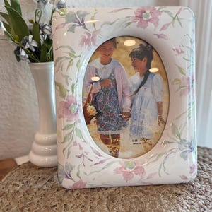 Puede incluir: Marco de fotos rectangular de cerámica blanca con apertura ovalada. Patrón floral en rosa, morado y verde. La foto muestra a dos niñas. Un jarrón blanco con flores a la izquierda.