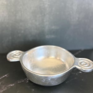 May include: A small, silver-colored bowl with two handles featuring a spiral design. The bowl has a rounded shape and a smooth, reflective surface. The handles extend outward from the sides.