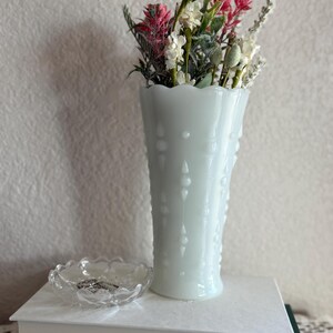 May include: A tall, light blue hobnail vase filled with a bouquet of dried flowers in shades of white, pink, and green. A small, clear glass dish sits on a stack of books next to the vase. The vase has a scalloped rim.