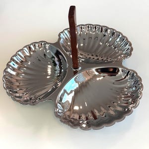 May include: A three-section serving dish featuring a polished silver-tone finish and a shell-shaped design for each compartment. A vertical, dark brown wooden handle is attached to the center, providing a grip for carrying the tray.