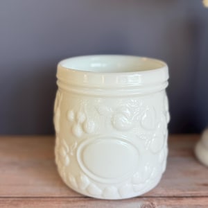 Puede incluir: Tarro de estilo vintage, color crema, con diseños florales y de bayas en relieve. El tarro tiene una boca ancha y un borde de rosca. Un espacio ovalado está centrado en la parte delantera. Hecho de vidrio opaco.