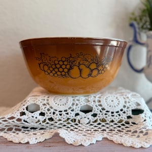 Puede incluir: Un cuenco mezclador Pyrex vintage con un degradado marrón y naranja. El cuenco presenta un diseño de fruta negra de uvas y peras. El cuenco está sobre un mantelito de encaje blanco.