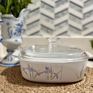 May include: White casserole dish with a clear glass lid. The dish features a floral design of purple irises and green stems. The dish is sitting on a woven placemat, with a blue and white pitcher in the background.