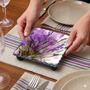 May include: A square decorative serving tray with a floral design featuring purple lavender flowers. The tray is made of slate and has small black feet. The background includes a table setting with plates, silverware, and a striped runner.