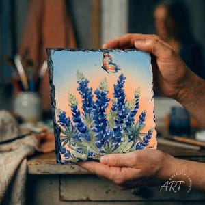 May include: A square piece of art featuring a watercolor painting of bluebonnets and a butterfly against a sunset sky. The artwork is on a dark, textured surface, held by hands. The bottom right corner has the text "ART".