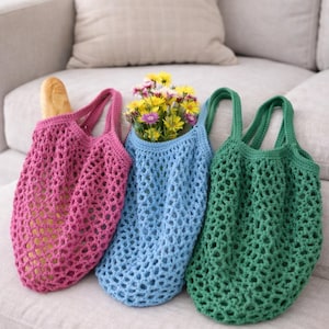 Könnte beinhalten: Drei gehäkelte Marktbeutel in Rosa, Blau und Grün liegen auf einem neutralfarbenen Sofa. Die rosa Tasche enthält ein Brot, die blaue Tasche enthält gelbe Blumen und die grüne Tasche ist leer. Die Taschen haben offene, netzartige Designs.
