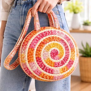 May include: A round handbag with a spiral design in shades of pink, orange, and cream. The bag has a matching strap and handles. The bag is made of woven material, and is a summer accessory.