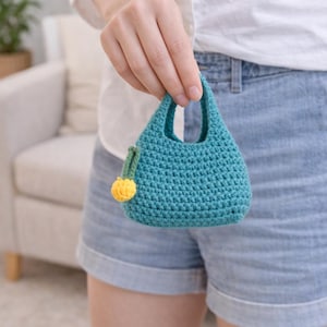 May include: Teal crochet handbag with a small handle and a yellow pom-pom accent. The handmade bag is a small, stylish accessory, perfect for carrying essentials. The bag is approximately 5 inches tall.