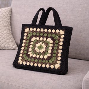 Könnte beinhalten: Eine schwarze Häkeltasche mit quadratischem Granny-Square-Design in Grün-, Braun- und Cremetönen. Die Tasche hat zwei schwarze Griffe und liegt auf einem grauen Sofa. Ein dekoratives Kissen befindet sich im Hintergrund.
