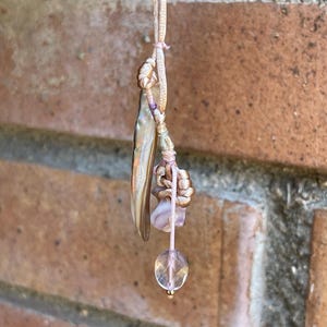 Könnte beinhalten: Eine Anhängerkette mit einem langen, schillernden, gebogenen Muschelstück, das an einer hellrosa Kordel hängt. Das Design umfasst einen kleinen, geknoteten rosa Stoffakzent, einen glatten lavendelfarbenen Stein und eine klare, facettierte Perle.