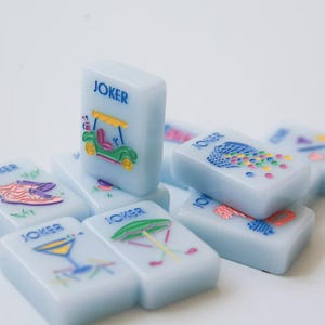 May include: A collection of light blue Mahjong tiles with colorful illustrations. Each tile is rectangular and features the word "JOKER" in blue. The images include a golf cart, a cocktail, and other playful designs. The tiles are arranged on a white surface.
