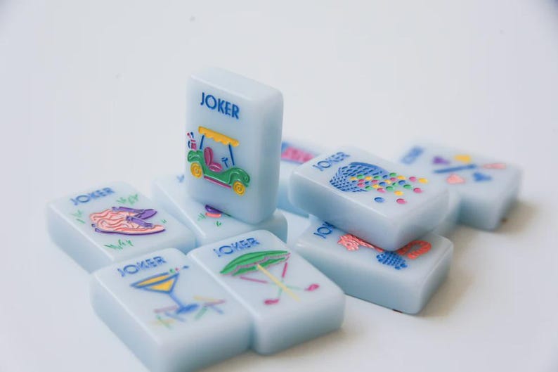 May include: A collection of light blue Mahjong tiles with colorful illustrations. Each tile is rectangular and features the word "JOKER" in blue. The images include a golf cart, a cocktail, and other playful designs. The tiles are arranged on a white surface.