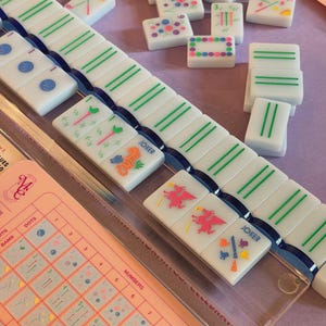 May include: A Mahjong game set with white tiles featuring green, blue, and pink designs. The set includes a pink game board with a clear plastic cover and a variety of tiles with different symbols and numbers.