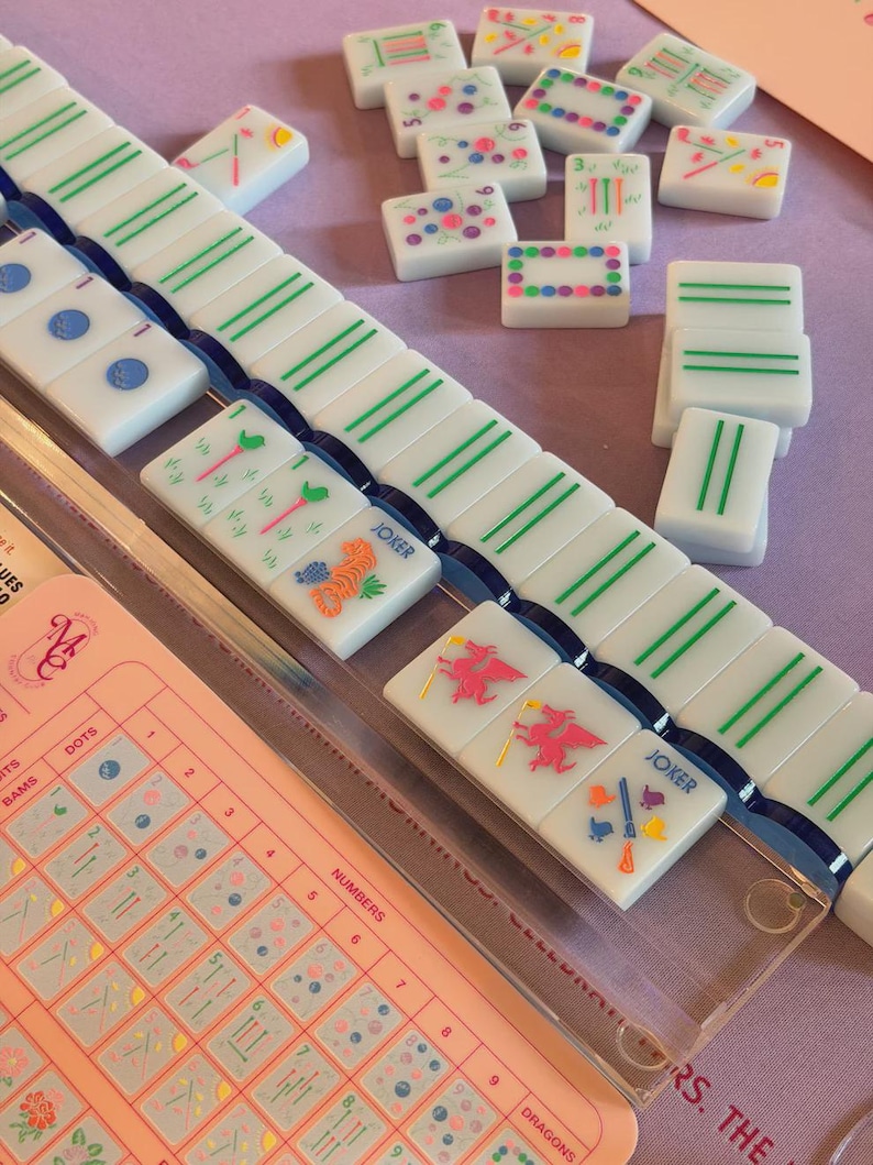 May include: A Mahjong game set with white tiles featuring green, blue, and pink designs. The set includes a pink game board with a clear plastic cover and a variety of tiles with different symbols and numbers.
