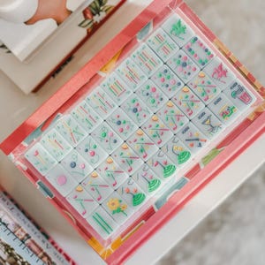 May include: A boxed set of Mahjong tiles with colorful illustrations. The tiles are white with various symbols in green, pink, yellow, and blue. The box is pink with a floral design and is sitting on a white surface.