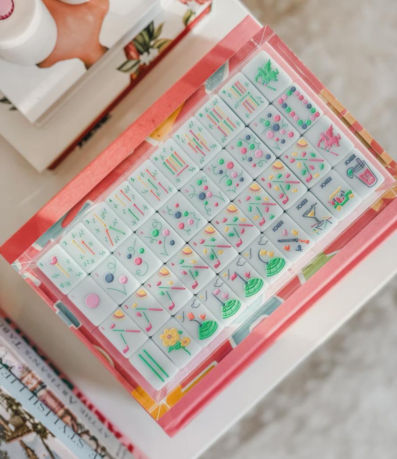 May include: A boxed set of Mahjong tiles with colorful illustrations. The tiles are white with various symbols in green, pink, yellow, and blue. The box is pink with a floral design and is sitting on a white surface.