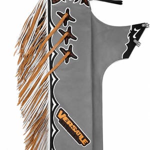 Op de afbeelding: Een grijze leren chaps-broek met lange witte en lichtbruine franjes aan de zijkant. Het kledingstuk is voorzien van bruine leren veters, zwarte randen met witte stiksels en een embleem met de tekst VERSATILE LEATHER in oranje en zwarte letters.