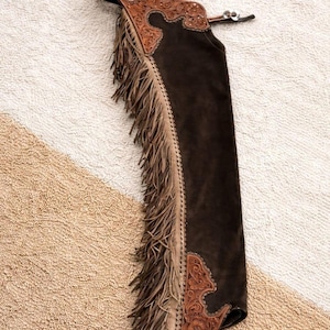 Op de afbeelding: Donkerbruine suède chaps met decoratieve lederen accenten en franjes. De chaps hebben ingewikkelde bloemen- en krulontwerpen, een zilverkleurige gesp en een gedetailleerde leren tailleband. De franje loopt langs de zijkant van de chaps.