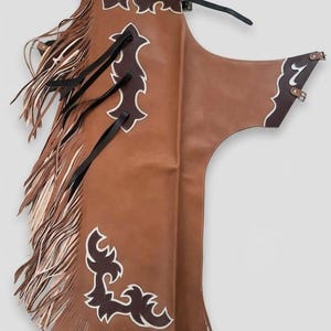 May include: Brown leather chaps with white and dark brown decorative accents. The chaps have a fringed edge and decorative cutouts. The belt and straps are black and brown.