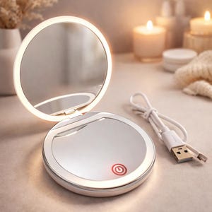 Könnte beinhalten: Ein silberner, runder, beleuchteter Schminkspiegel, der geöffnet ist, um eine reflektierende Oberfläche zu zeigen. Der Spiegel hat ein eingebautes LED-Licht am Rand. Ein weißes USB-Ladekabel ist angeschlossen. Der Power-Knopf leuchtet rot.