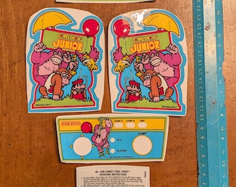 Op de afbeelding: Een verzameling Donkey Kong Junior-game-stickers en een sticker voor het bedieningspaneel. De stickers tonen het logo en de personages van de game, waaronder Donkey Kong en Junior. De sticker van het bedieningspaneel heeft knoppen en tekst.