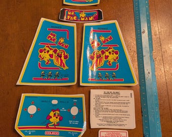 Op de afbeelding: Een verzameling Ms. Pac-Man arcade game onderdelen, waaronder bedieningspanelen en decoratieve panelen. De stukken hebben een blauwgroen en geel kleurenschema met het Ms. Pac-Man karakter. Een blauwe liniaal is inbegrepen.