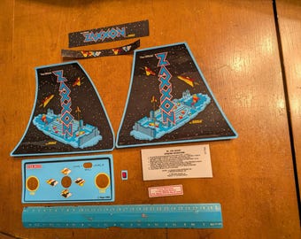 Op de afbeelding: Een verzameling vintage Zaxxon arcade game onderdelen, waaronder twee grote zijpanelen met een ruimtescène, een bedieningspaneel en diverse stickers. De onderdelen zijn blauw, zwart en rood, met het Zaxxon logo.
