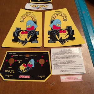 Op de afbeelding: Een verzameling vintage Pac-Man arcade game onderdelen. Inclusief gele en zwarte bedieningspaneel overlays, kastkunst met het Pac-Man spookkarakter en een Midway logo. Bevat ook bedieningsinstructies.