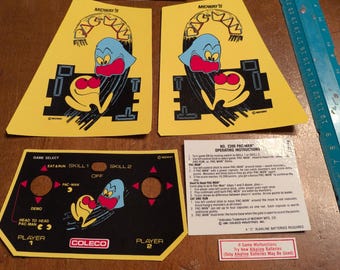 Op de afbeelding: Een verzameling vintage Pac-Man arcade game onderdelen. Inclusief gele en zwarte bedieningspaneel overlays, kastkunst met het Pac-Man spookkarakter en een Midway logo. Bevat ook bedieningsinstructies.