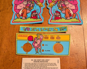 Op de afbeelding: Een verzameling Donkey Kong Junior-game-stickers en instructies. De stickers tonen personages als Donkey Kong, Mario en vogels. De instructies bevatten bedieningsdetails en copyrightinformatie. De kleuren zijn levendig, met blauw, rood en geel.