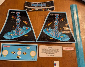 Op de afbeelding: Een verzameling vintage arcade game stickers en onderdelen. Inclusief twee driehoekige stickers met "ZAXXON" tekst en een ruimtescène, een sticker voor het bedieningspaneel en andere gerelateerde onderdelen. Het Sega logo is zichtbaar.
