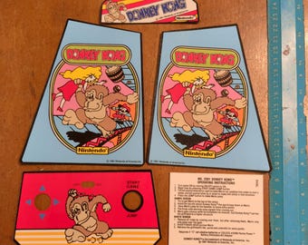 Op de afbeelding: Een verzameling Donkey Kong arcade game stickers en instructies. De stickers tonen Donkey Kong, Mario en Pauline. De kleuren zijn blauw, rood, geel en bruin. De instructies zijn in het Engels.