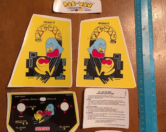 Op de afbeelding: Een verzameling Pac-Man arcade game stickers en bedieningspaneel overlays. De stickers tonen het Pac-Man karakter in verschillende ontwerpen, met de Midway en Coleco logo's. Een blauwe liniaal is inbegrepen voor de schaal.