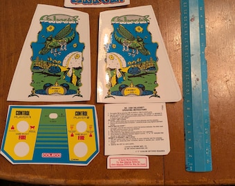 Op de afbeelding: Een verzameling vintage arcade game stickers en instructies. Bevat twee grote zijstukken met een groen insect en ruimteschip ontwerp, een bedieningspaneel en een liniaal tot 30 cm.