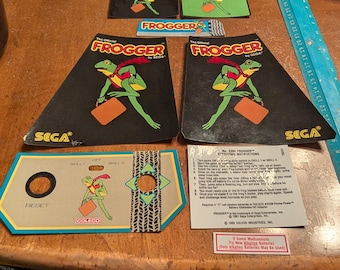 Op de afbeelding: Een verzameling vintage Frogger-gamecomponenten, waaronder instructiebladen, bedieningspanelen en game-art. De afbeelding toont een groene kikker met een bruine aktetas. De logo's van Sega en Coleco zijn zichtbaar.