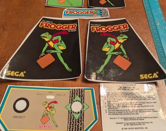 Op de afbeelding: Een verzameling vintage Frogger arcade game stickers en instructiebladen. De stickers tonen een groen kikkerkarakter met een rode sjaal en aktetas. De logo's van Sega en Coleco zijn zichtbaar. Inclusief een liniaal.
