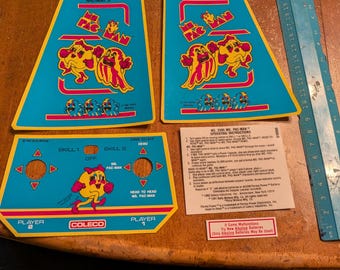 Op de afbeelding: Een verzameling vintage Ms. Pac-Man arcade game onderdelen. De set bevat blauwe en gele bedieningspaneel overlays, een marquee en een instructieblad. De graphics tonen het Ms. Pac-Man karakter en het Midway logo.