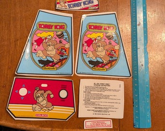 Op de afbeelding: Een verzameling vintage Donkey Kong arcade game onderdelen en stickers. Inclusief twee grote blauwe en gele kastpanelen, kleinere stickers en een rood bedieningspaneel. Er is ook een blauwe liniaal aanwezig.