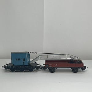 Puede incluir: Un juego de trenes modelo con un vagón grúa azul y un vagón plataforma rojo. El vagón grúa tiene una pluma plateada y el vagón plataforma tiene ruedas negras. Los vagones están conectados y se encuentran sobre una superficie blanca.