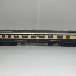 Può includere: Un vagone ferroviario modello con tetto argentato, finestre color crema e base blu scuro. Le parole "RHEINGOLD" sono stampate in lettere dorate sul lato. Il vagone è una replica in miniatura.